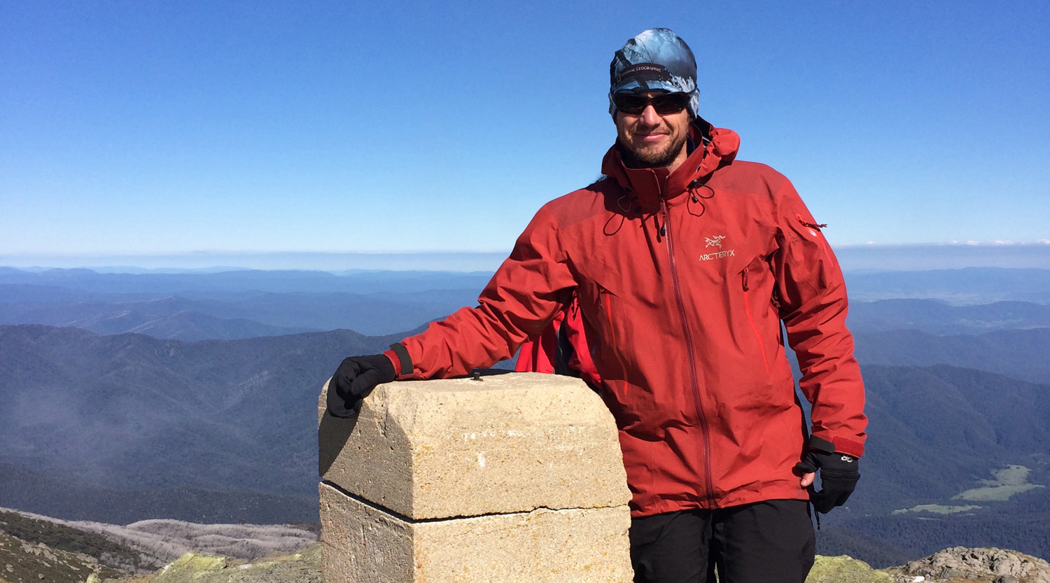 Brendon Munge, dressed in a red jacket, beanie, sunnies and gloves, on top of Mount Townsend in the Kosciuszko Range during an 18-day walk with university students