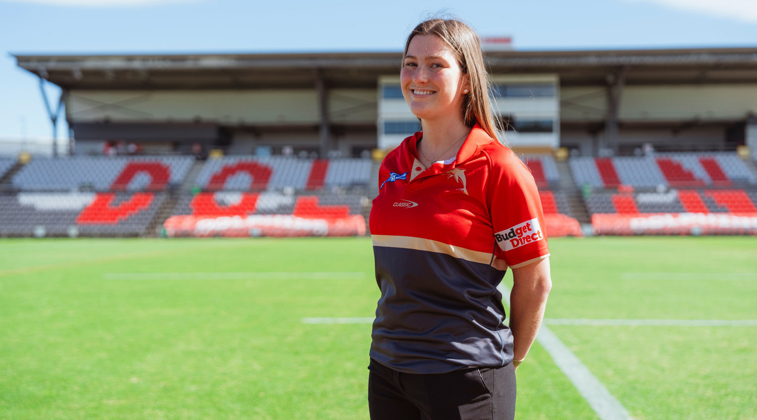 Emily Hill, wearing red and black Dolphins NRL colours, is standing next to a green footy field with stadium seating in the background that has Dolphins written across the seats