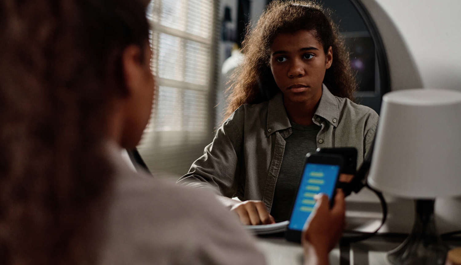 teenage girl with smartphone sitting in front of mirror and looking at her reflection while reading cyberbullying messages