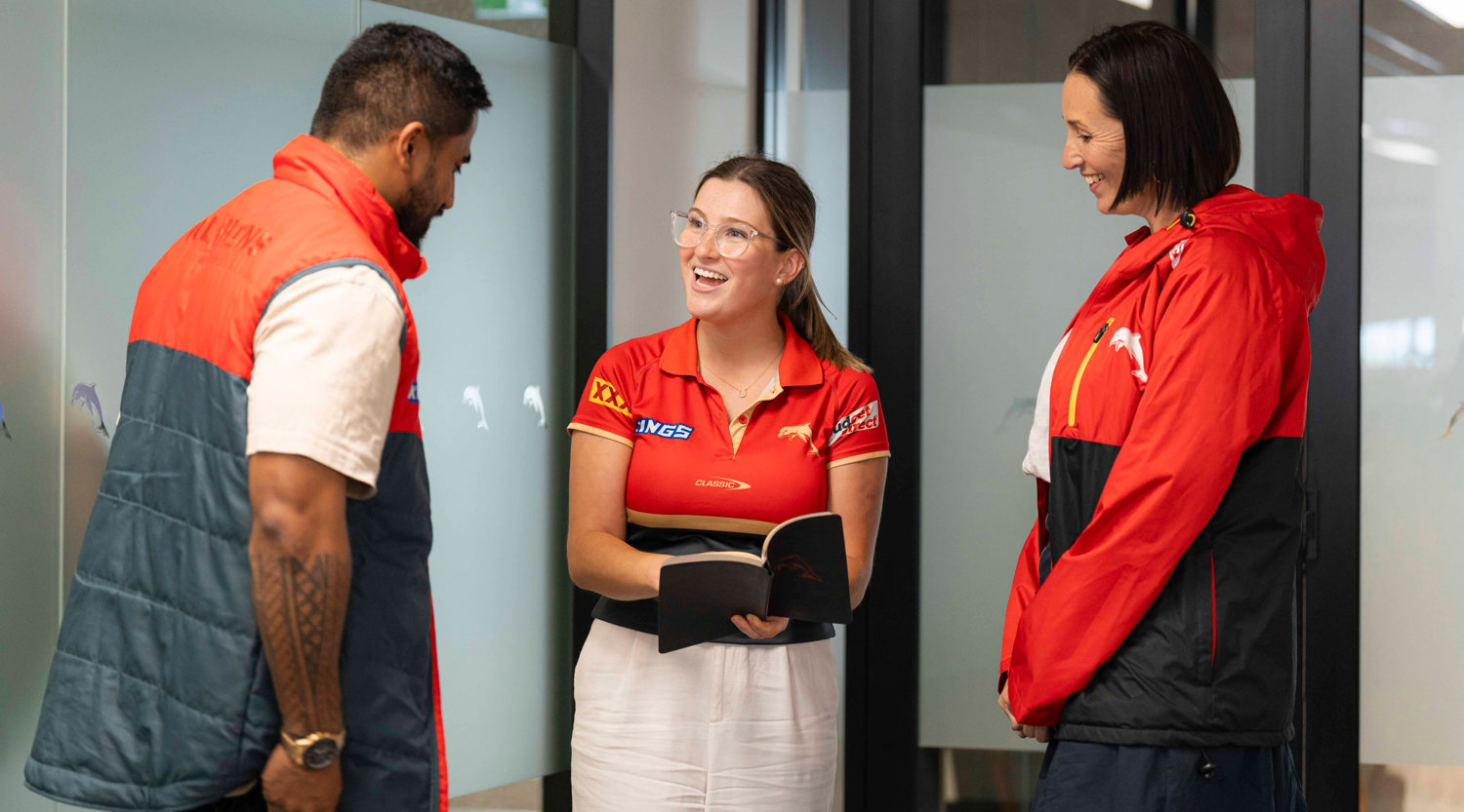 Emily Hill is standing talking with colleagues all dressed in red Dolphins uniforms in an office setting